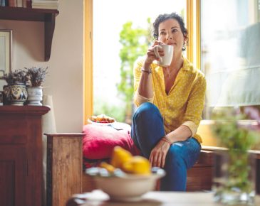 Pensive looking woman holding coffee cup to her lips while sitting in an opened window sill