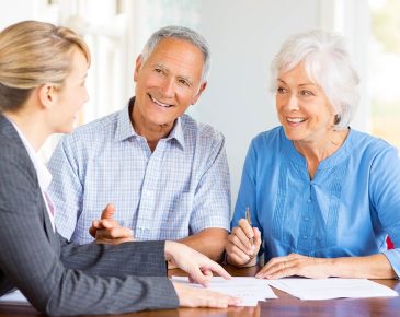 older couple smiling and speaking to a woman wearing a suit while pointing to papers on the table in front of them
