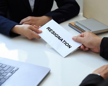 zoomed in perspective of two people sitting at a table exchanging a piece of paper that reads "resignation"