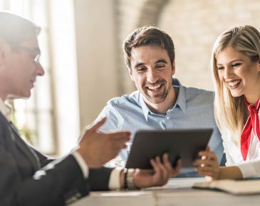 Smiling man and woman looking at computer tablet while a man in a suit explains something to them