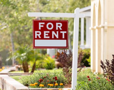 Red sign that reads "for rent" hanging from white post in landscaped front yard